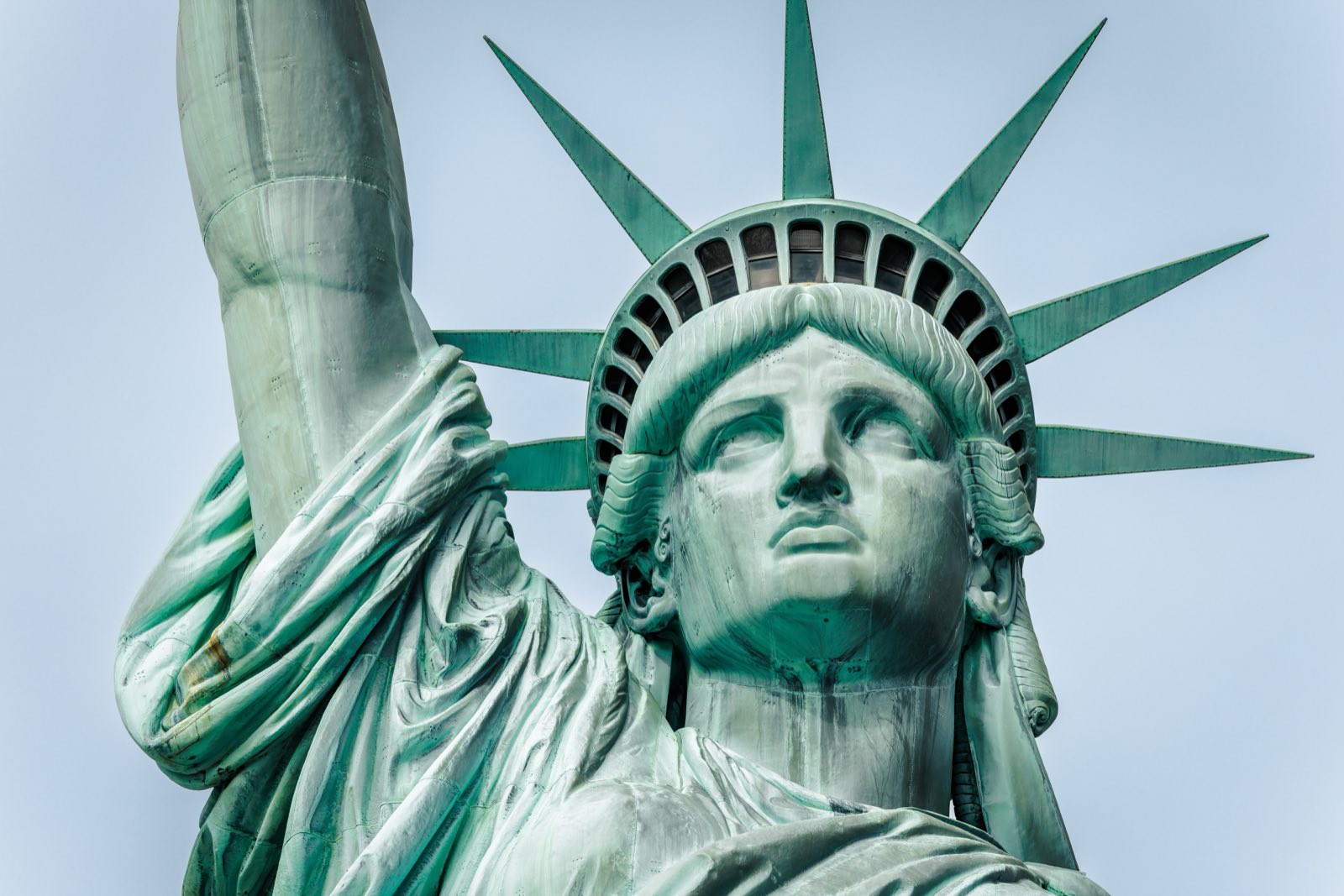 A close view of the Statue of Liberty's face and crown, her raised arm behind, against a clear pale sky